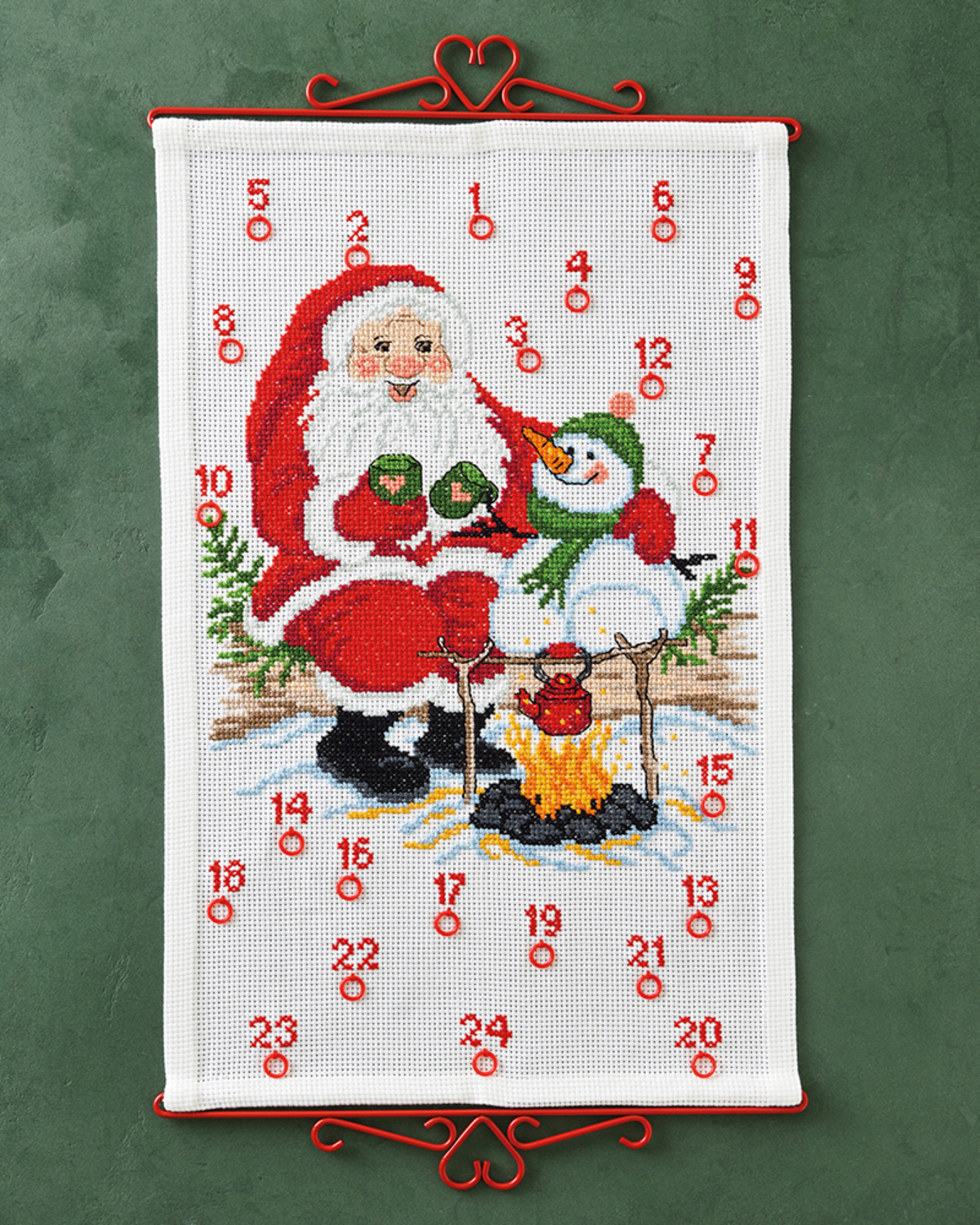 Broderet julekalender med julemand og snemand ved bålet, med ringe til 24 pakker