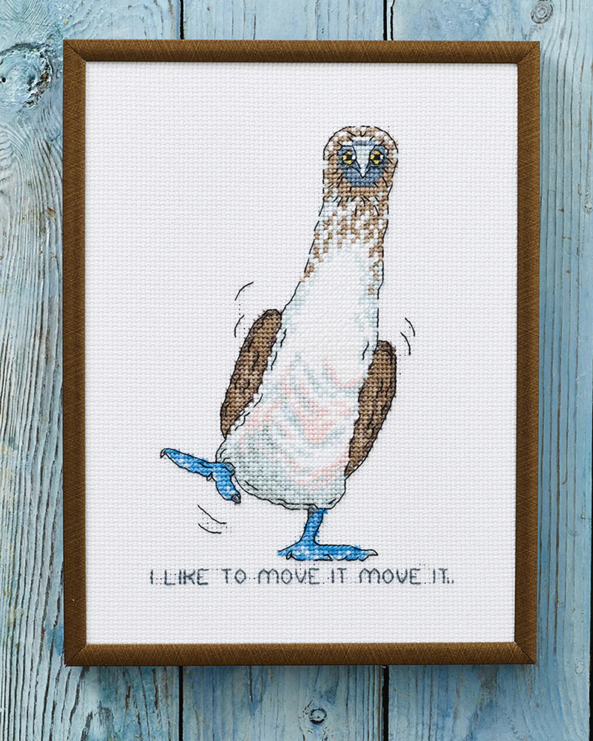 Broderikit i korssting med en fugl med blå fødder og teksten I like to move it move it på hvid Aida, i ramme